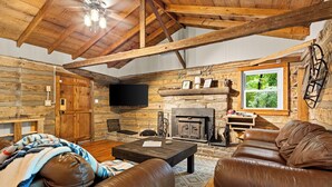 Interior rústico de una cabaña de troncos con chimenea, muebles de cuero y una televisión.