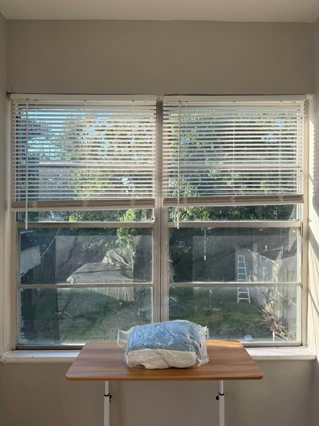 A room with a window overlooking a backyard. There is a wooden table with a plastic-wrapped item on it.
