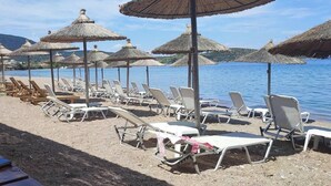 Een strand met rijen ligbedden en rieten parasols, een houten terras en uitzicht op de zee.
