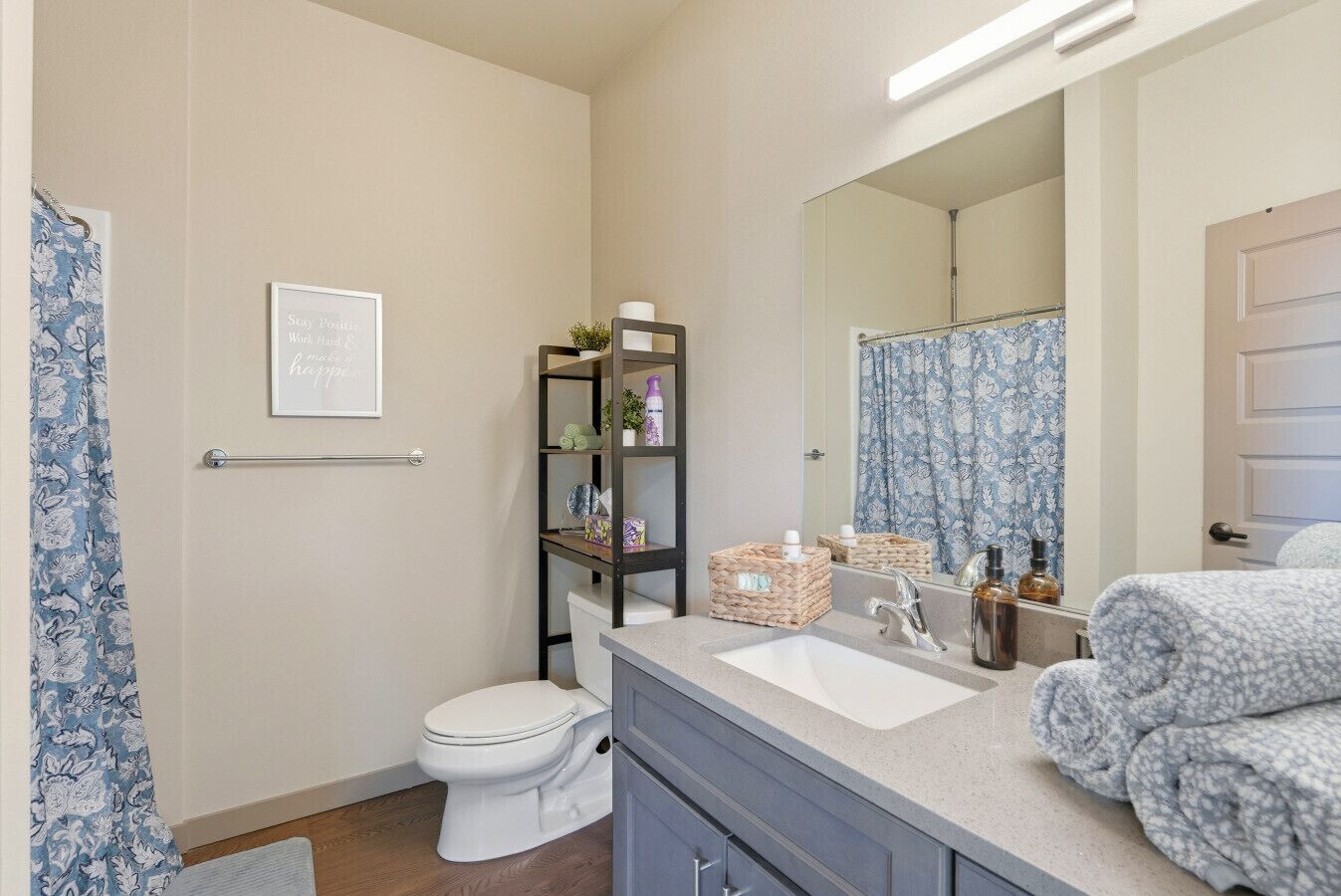 A bathroom with a toilet, sink, and mirror. There is a shelf with toiletries and a framed quote on the wall.