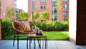 A wicker chair with cushions and a glass of wine on a round table, set on a patio with a brick building and greenery in the background. - City Center - Łąkowa street by Apartmore (Gdańsk)