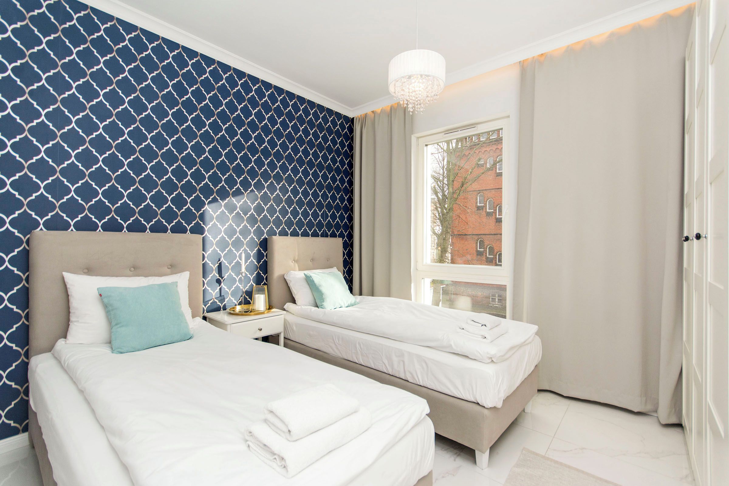 A hotel room with two beds, a large window with a view of a building, and a chandelier.