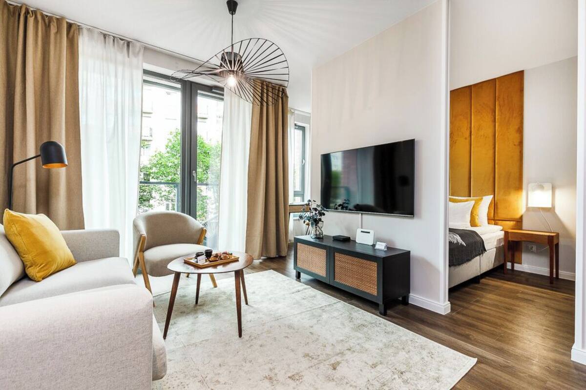 A modern living room with a sofa, armchair, coffee table, TV, and a bed with a nightstand.