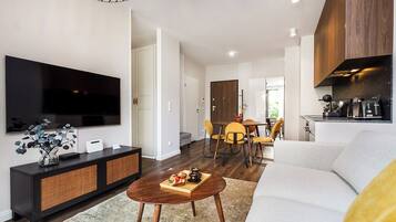 A modern living room with a flat-screen TV, a wooden coffee table with a tray of snacks, a sofa, and a dining area with yellow chairs.
