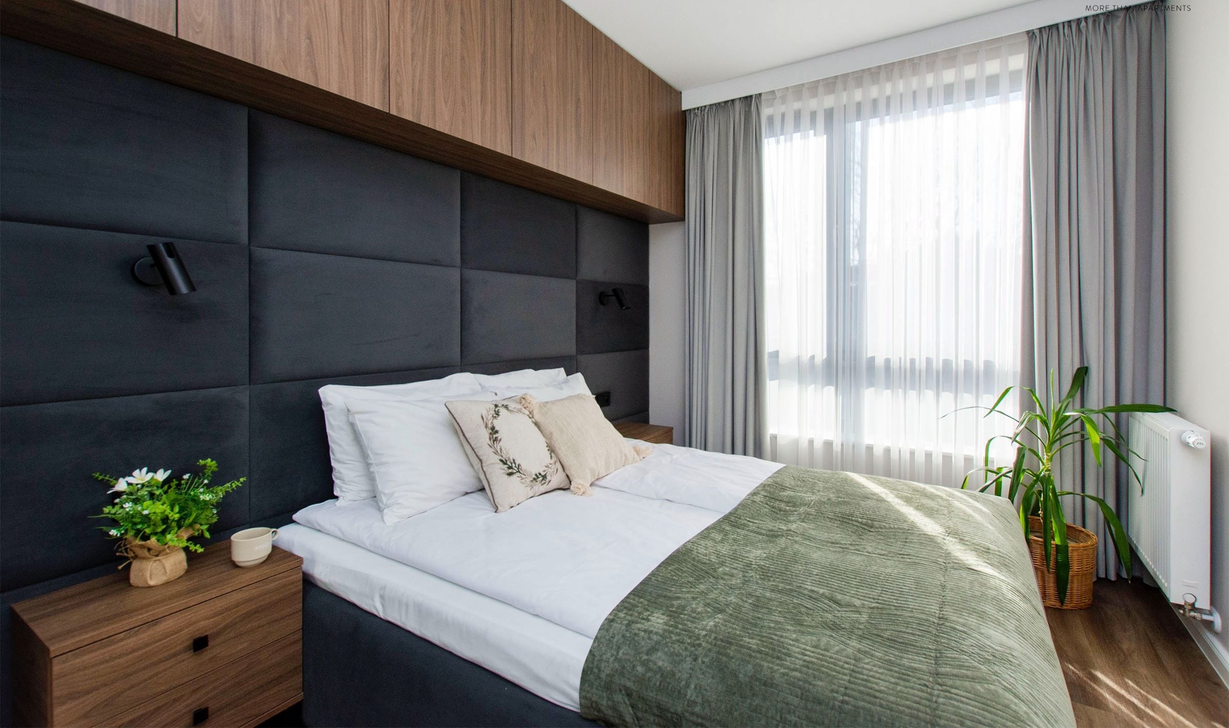 Une chambre à coucher moderne avec une tête de lit sombre, une table de chevet en bois, un plaid vert et une plante en pot.