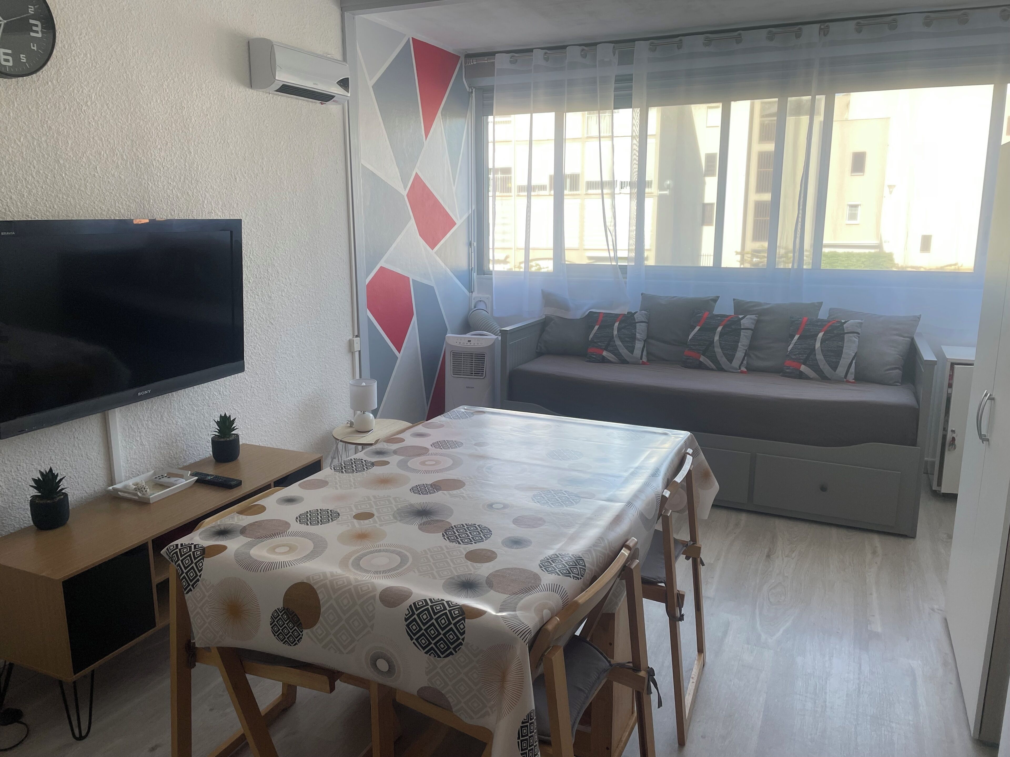 A living room with a TV, a dining table, a sofa, and a window with a view of buildings.