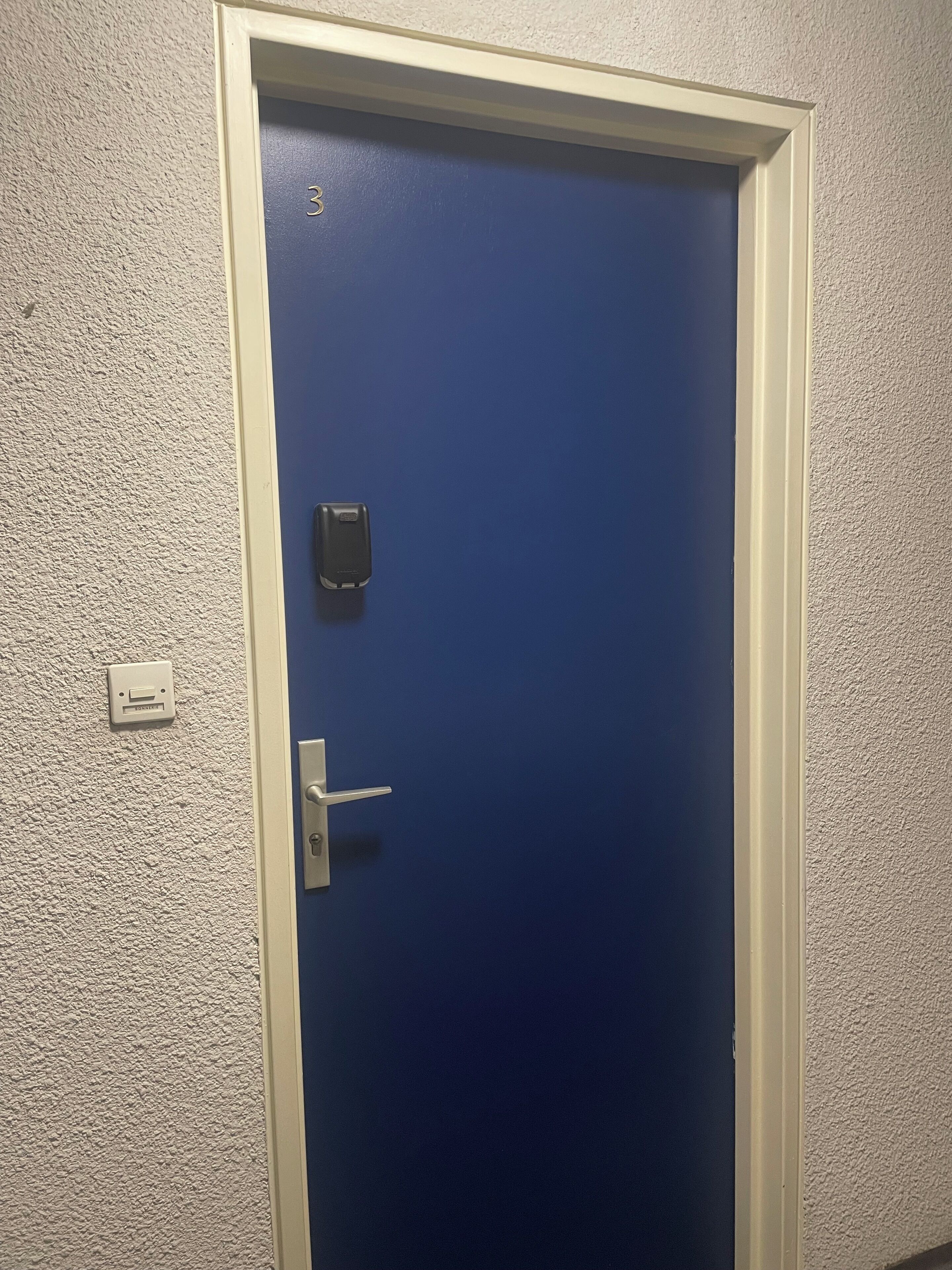En blå dør med hvid ramme, en ringeklokke og en intercom fastgjort på væggen.