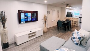 Una sala moderna con un televisor de pantalla plana montado en la pared, un sofá gris con cojines estampados, un mueble blanco para entretenimiento y un comedor al fondo.