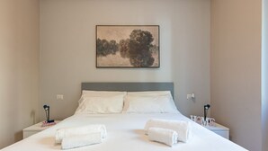 Un letto rifatto con lenzuola bianche e due asciugamani, un quadro appeso al muro e un comodino con una lampada e un cartello con scritto 