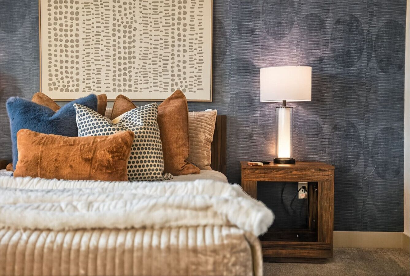 Un dormitorio ordenado con cama, almohadas, una mesita de noche con lámpara y una obra de arte en la pared.