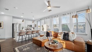 A modern living room with a brown leather sofa, a round coffee table, and a dining area with bar stools. - Rhythm Room | Oak Lawn 2BR Near Uptown, Pool & Gym (Dallas)