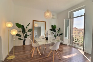 Ένας χώρος τραπεζαρίας με ένα τραπέζι στρωμένο για τέσσερα άτομα, έναν μεγάλο καθρέφτη και θέα μέσα από ένα ανοιχτό παράθυρο.