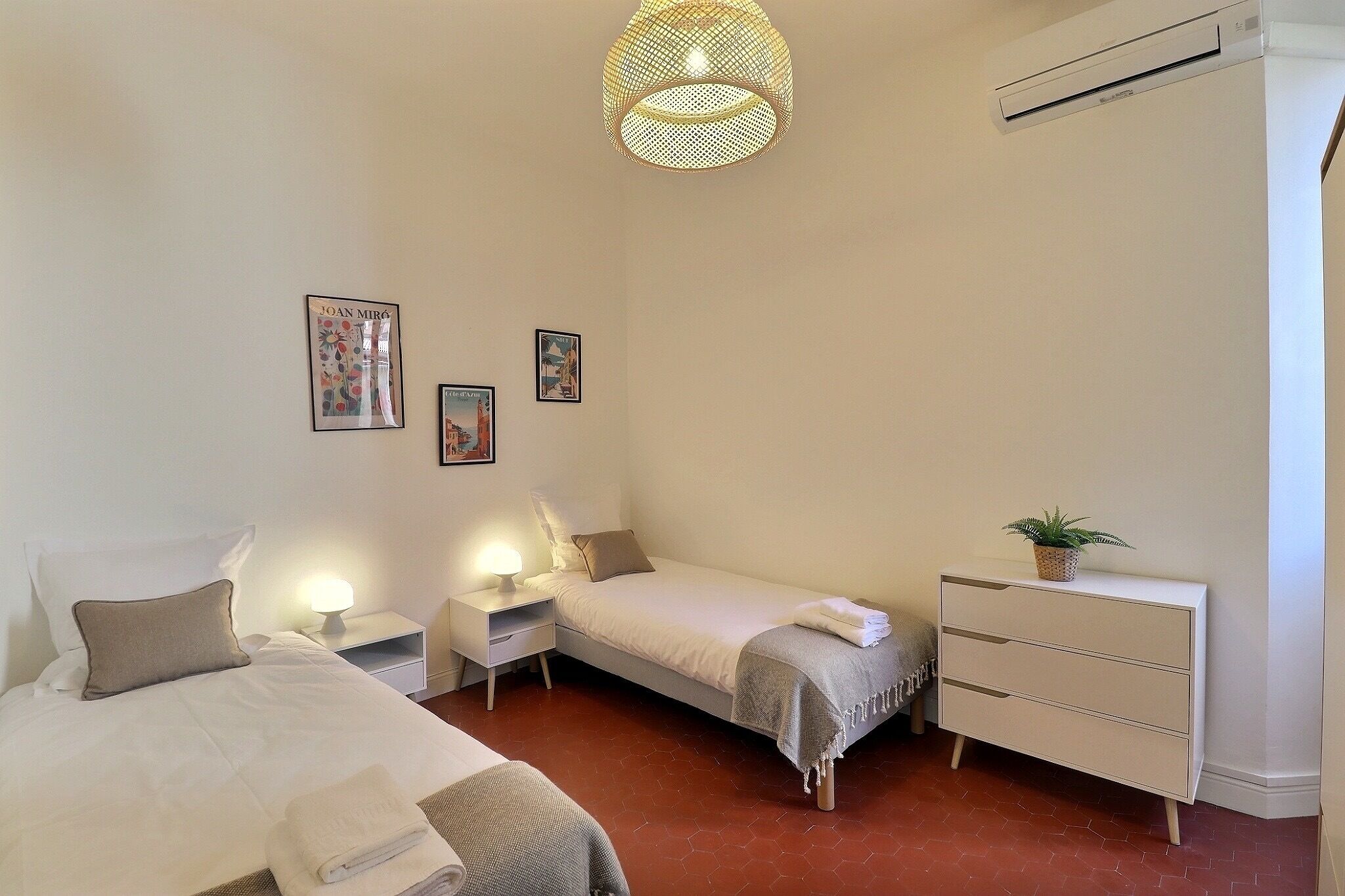 A bedroom with two beds, a bedside table with a lamp, a dresser, and wall art.
