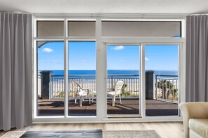 A room with a large sliding glass door leading to a balcony with two white chairs and a view of the ocean.