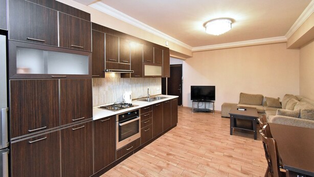 Una cucina moderna con mobili in legno scuro, forno integrato e una grande isola con lavello. Nella zona living, si trova una TV a schermo piatto e un divano.