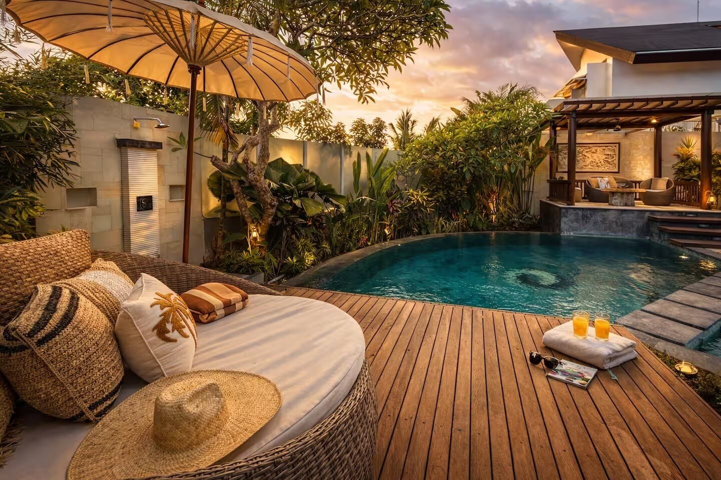 Une terrasse en bois avec une piscine, un canapé en osier agrémenté de coussins, un chapeau de paille et une table avec des boissons.