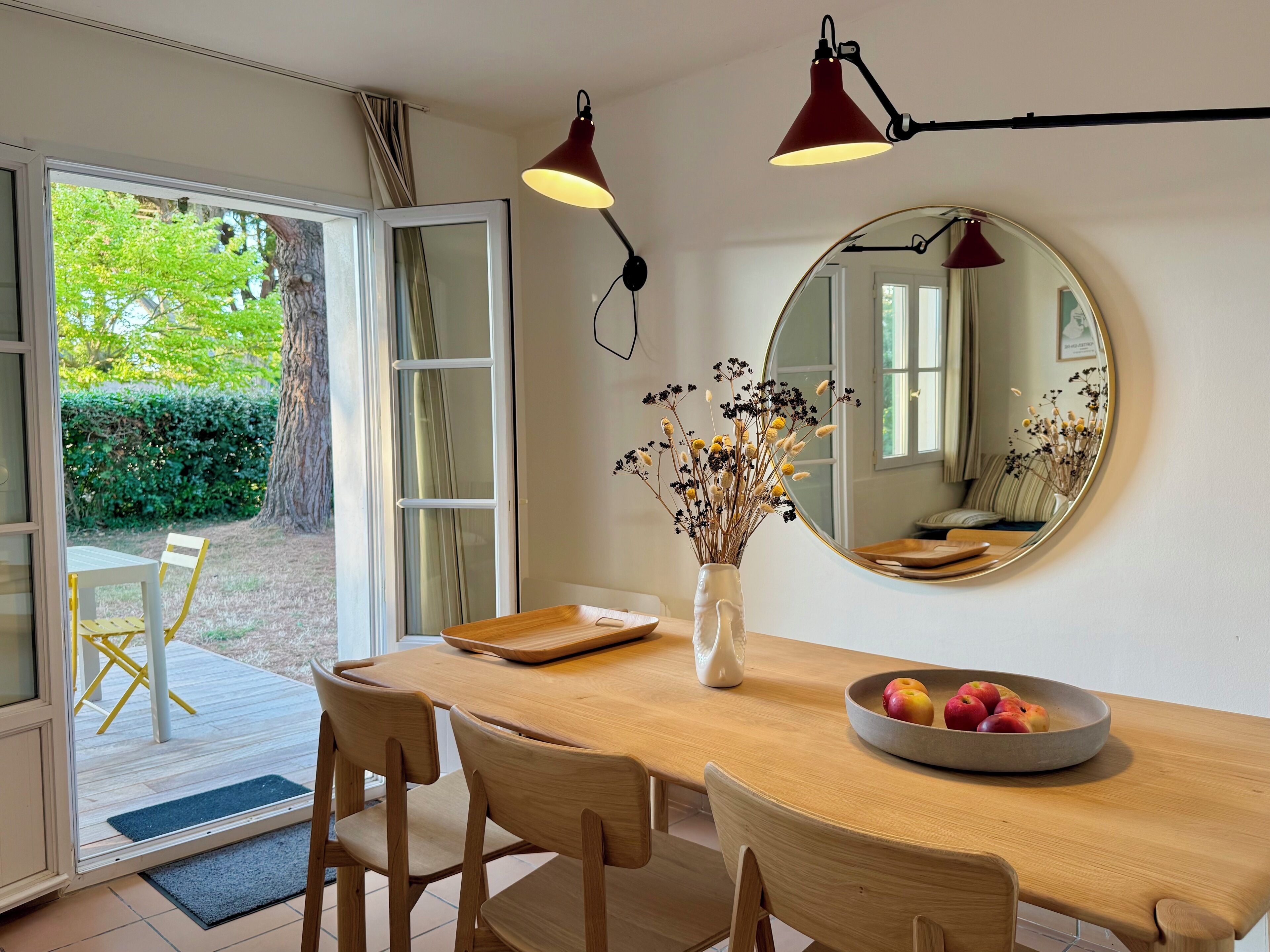 A dining area with a wooden table, chairs, a vase with dried flowers, a mirror, and a bowl of fruit.