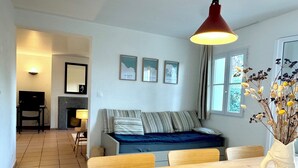A modern living room with a sofa, a dining table with a fruit bowl, and a vase with dried flowers. There is a fireplace, a lamp, and framed pictures on the wall. - La Maison de Vacances 3 minutes from the ocean (La Flotte)