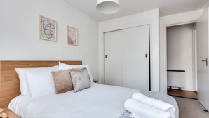 Een net opgemaakt bed met witte lakens en een houten hoofdbord, een witte plafondlamp en twee ingelijste kunstwerken aan de muur.