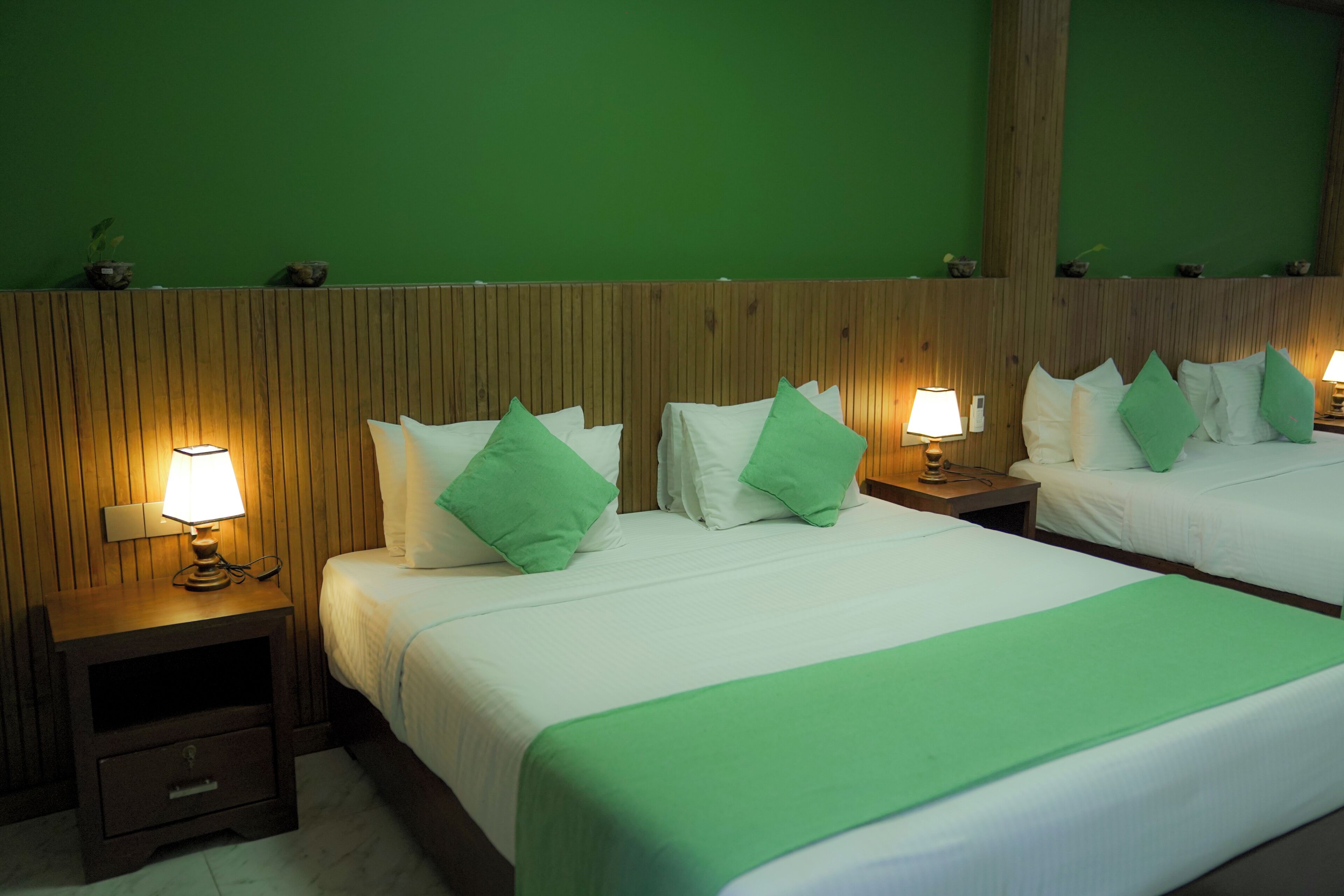 Habitación de hotel con dos camas, cada una con ropa de cama blanca y almohadas verdes, cabeceros de madera y lámparas de noche.