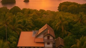 A two-story house with a red roof, surrounded by lush greenery and palm trees, overlooking a calm lake at sunset.