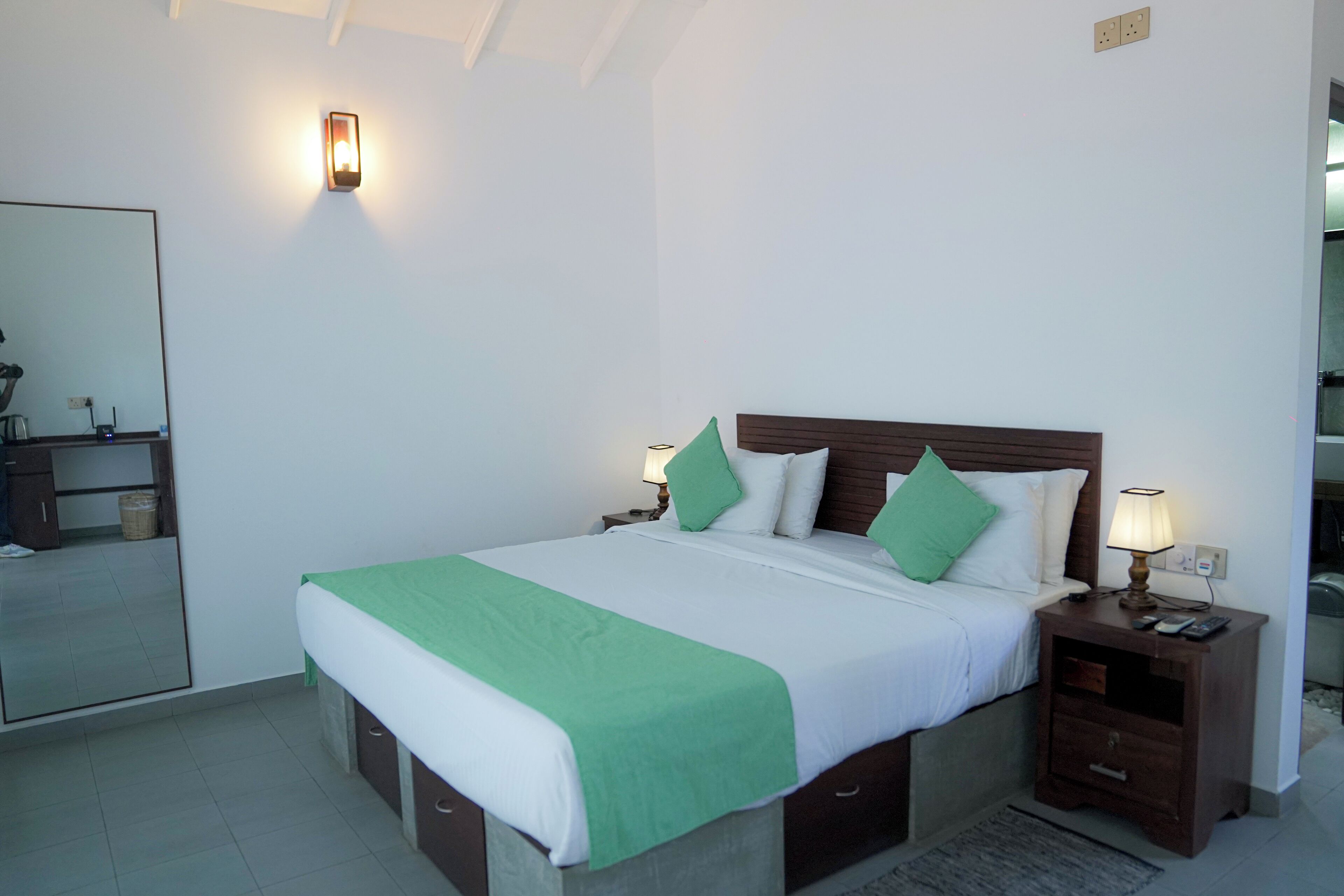 Een net opgemaakt bed met groen en witte beddengoed, een nachtkastje met een lamp en een spiegel aan de muur.