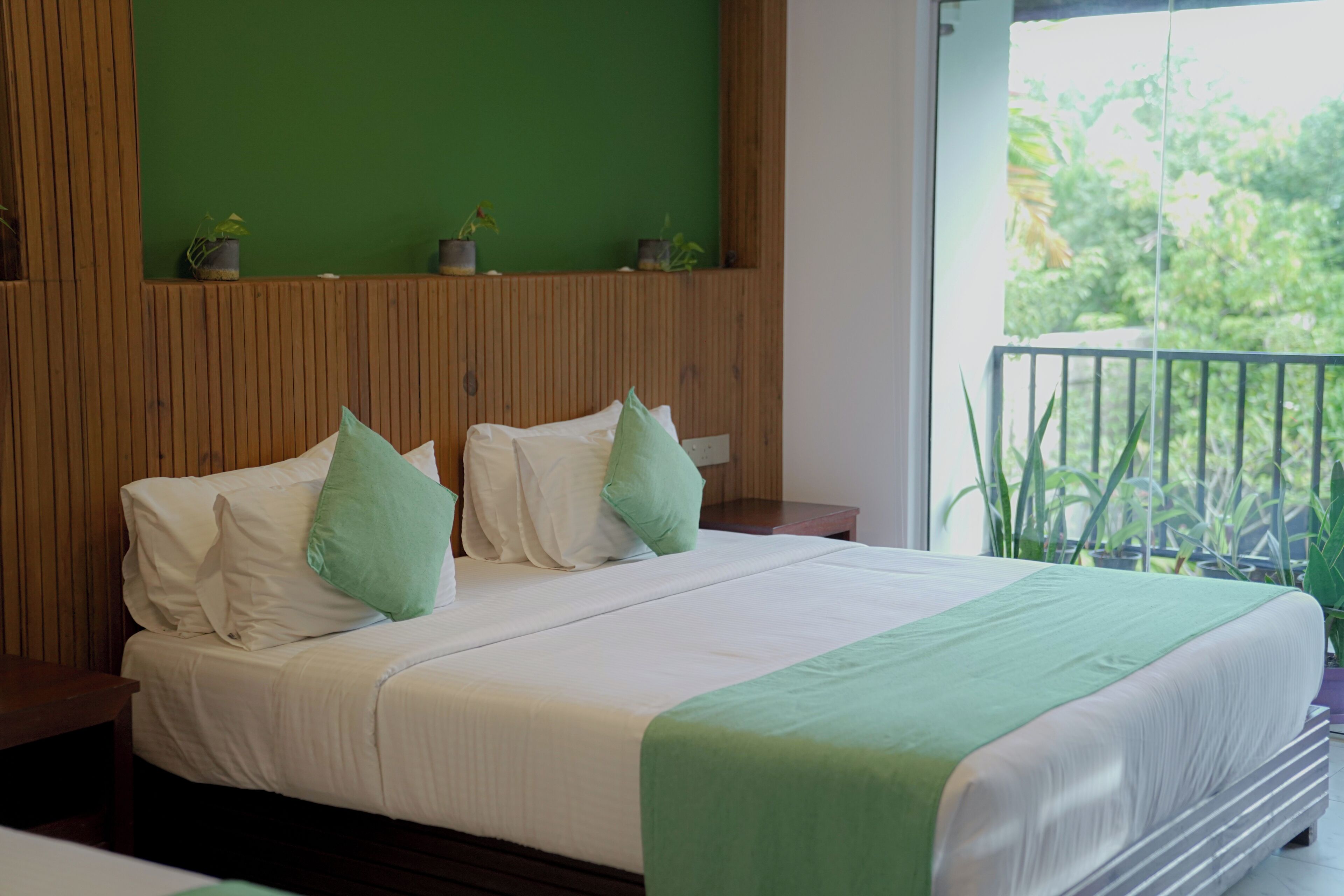 Cama con sábanas blancas, almohadas verdes, cabecera de madera y vista a plantas desde un balcón.