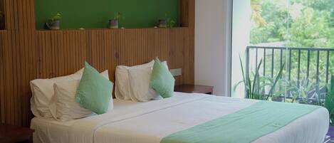 Cama con sábanas blancas, almohadas verdes, cabecera de madera y vista a plantas desde un balcón.