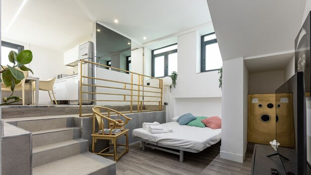 Ett modernt och kompakt bostadsutrymme med en trappa, en säng, en liten sittgrupp och ett litet kök.