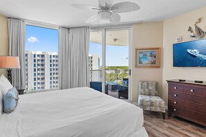 Quarto de hotel com uma cama grande, uma cadeira, cômoda, TV e varanda com vista para edifícios e vegetação.