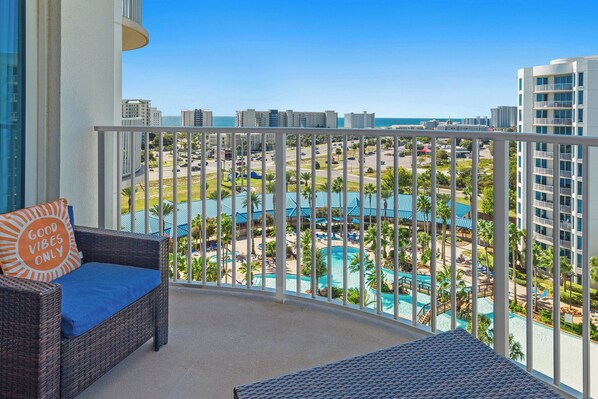 Varanda com sofá de vime e almofada, com vista para área de resort, piscina, palmeiras e oceano.