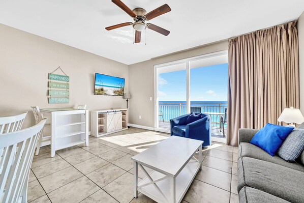 A spacious living room with a large window, a ceiling fan, a white coffee table, a grey sofa, and a blue armchair.