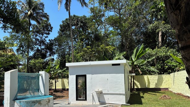 Pequeño edificio de una sola planta, blanco, con una puerta y una ventana, rodeado de abundante vegetación y un césped cuidado.