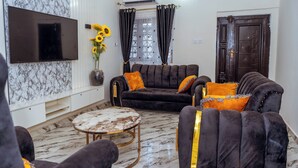A living room with a sofa, armchairs, a coffee table, a TV, and a window with curtains.