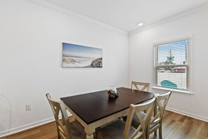 A dining area with a wooden table and chairs, a painting on the wall, and a window with blinds.