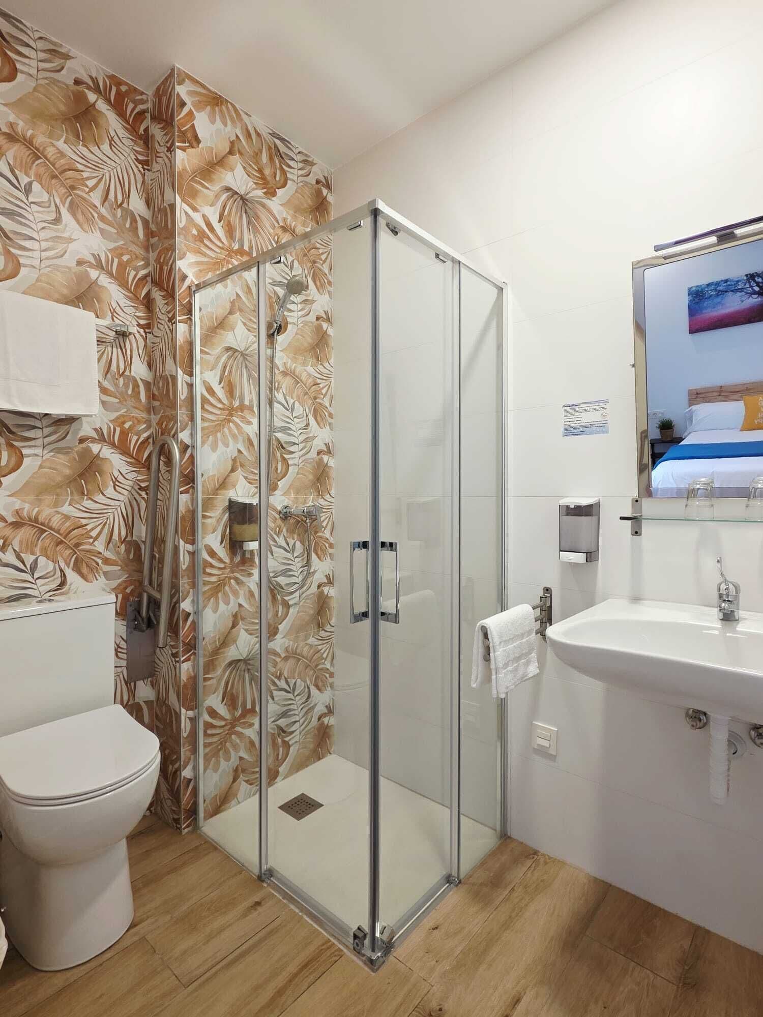 Casa de banho com cabine de chuveiro em vidro, sanita, lavatório com espelho e papel de parede com padrão floral.