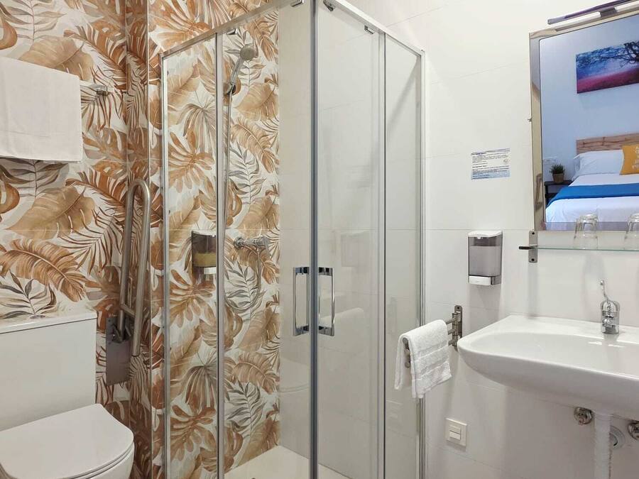 Casa de banho com cabine de chuveiro em vidro, sanita, lavatório com espelho e papel de parede com padrão floral.
