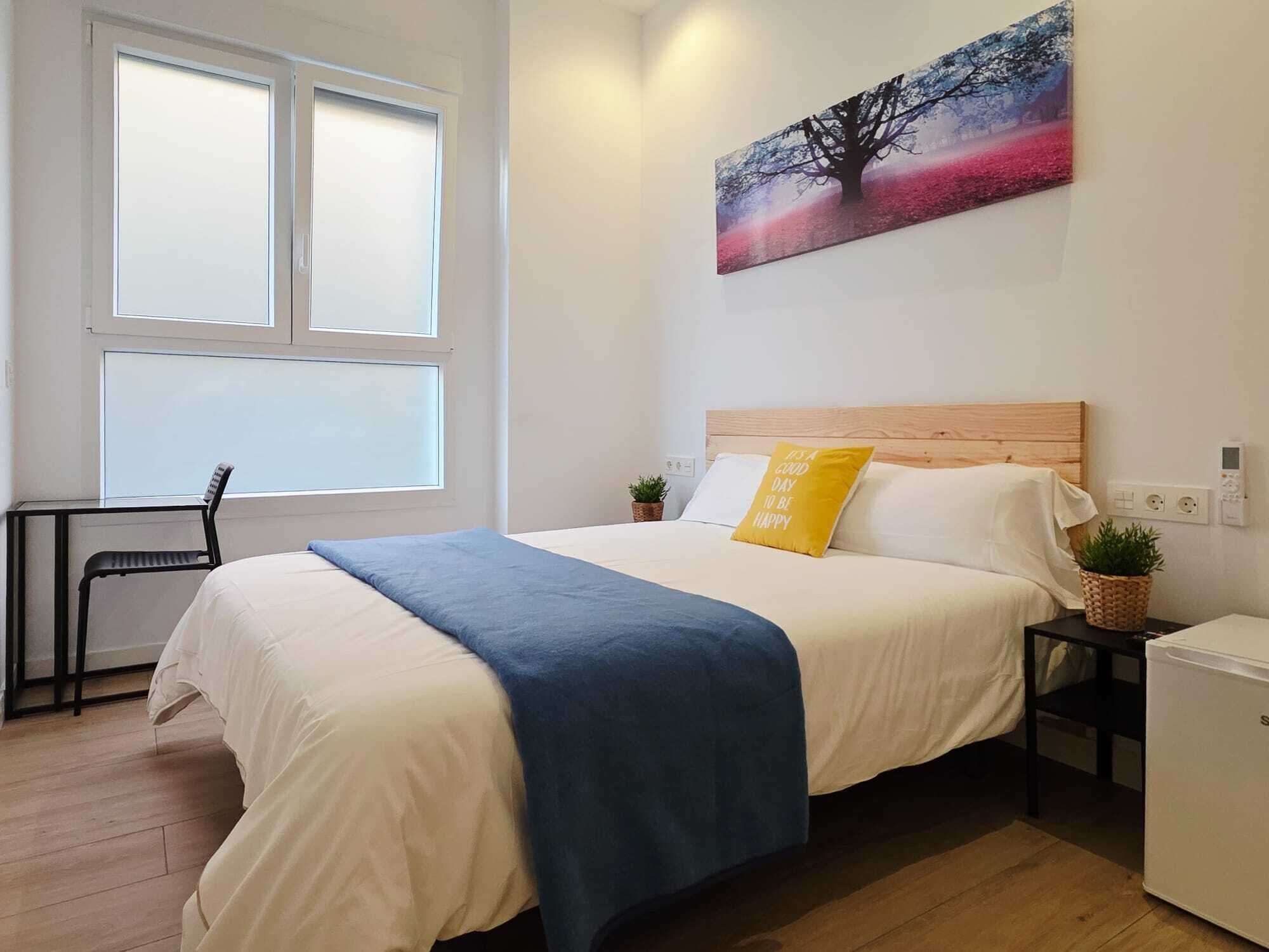 A bedroom with a bed, a desk, a chair, a small table with a plant, and a wall-mounted painting.