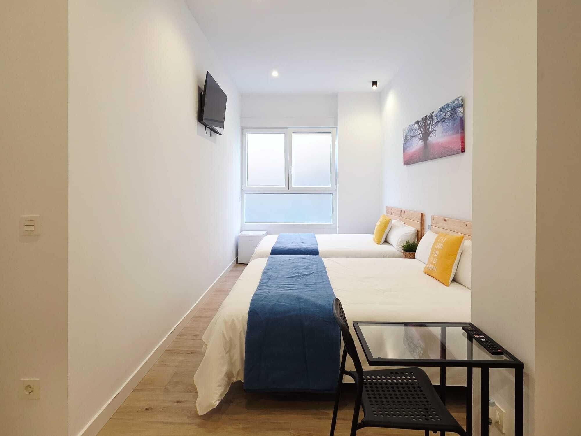 Habitación de hotel con dos camas, un televisor montado en la pared, una mesita y una ventana con persianas.