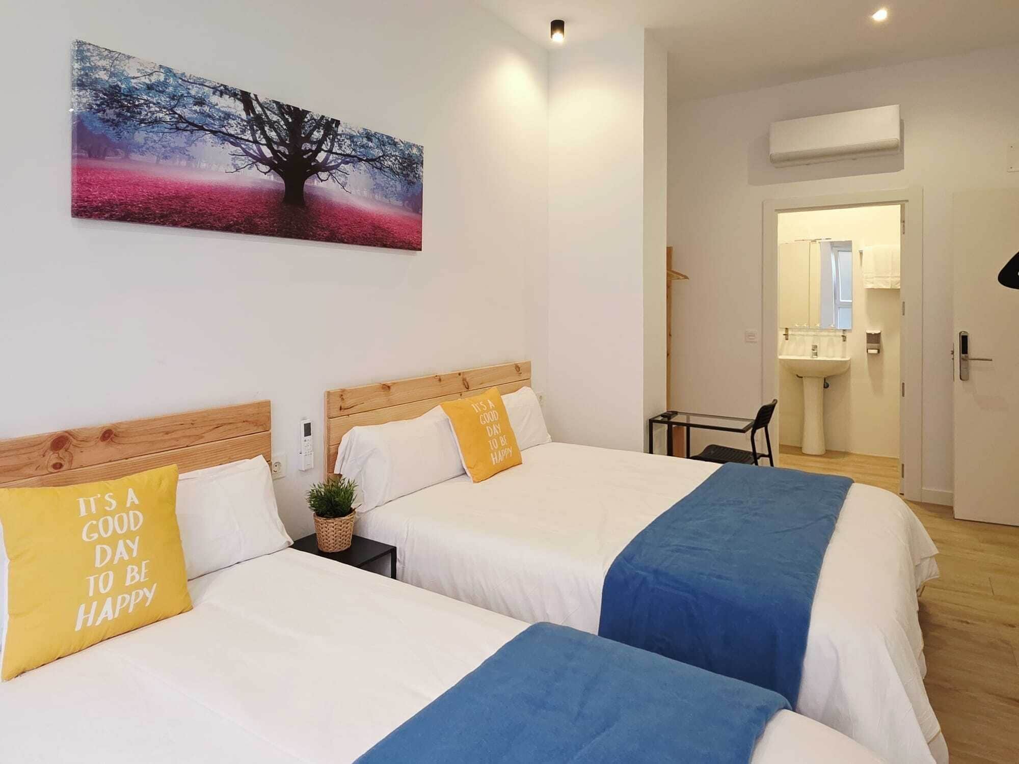 Camera d'albergo con due letti, un quadro raffigurante un albero, un tavolino e un bagno visibile attraverso una porta aperta.