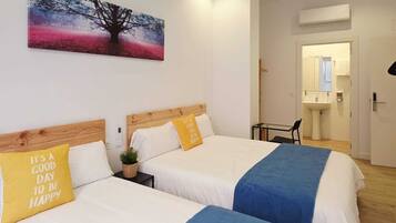 Habitación de hotel con dos camas, un cuadro de un árbol, una mesita y un baño visible a través de una puerta abierta.