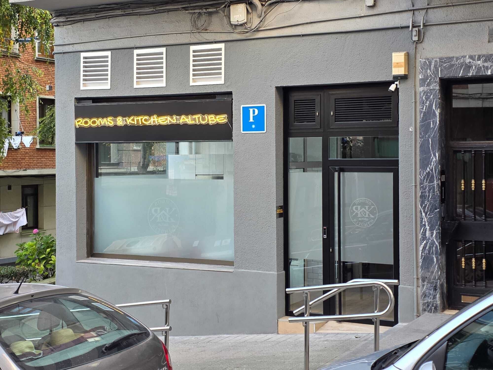Un edificio con un cartel que dice 'ROOMS & KITCHEN ALTUBE', un cartel de estacionamiento y una entrada con puerta de vidrio.