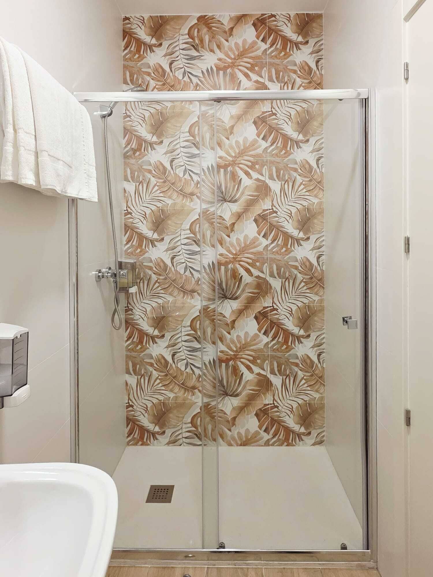 A glass-enclosed shower with a leaf-patterned wall, a white bathtub, and a towel on the wall.