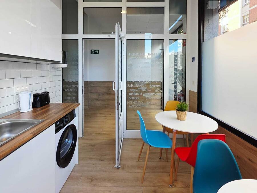 Una kitchenette compacta con lavadora, cafetera y fregadero. Hay una mesa pequeña con sillas y una planta en maceta.