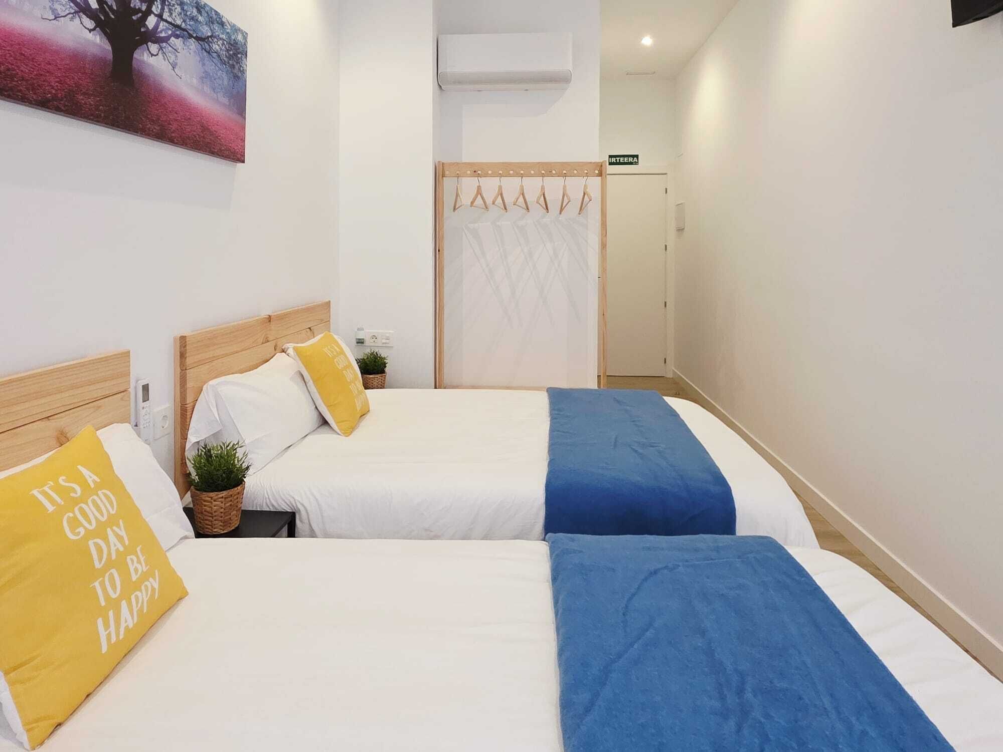 Hotellihuone, jossa on kaksi sänkyä, pieni ruukkukasvi, seinälle asennettu ilmastointilaite ja ovi, jossa on kyltti, jossa lukee ”IRTEERA”.