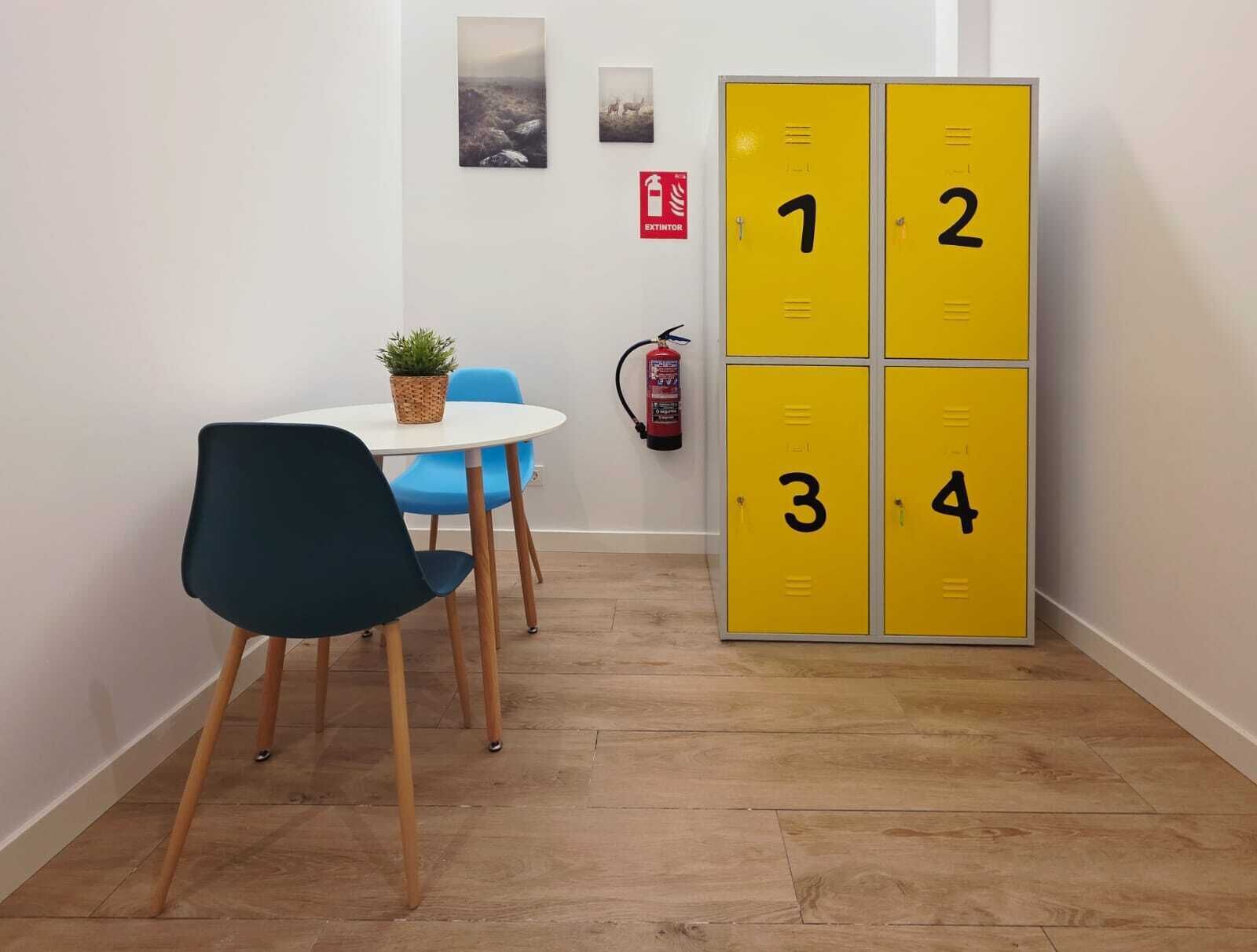 Una habitación con una mesa blanca, sillas azules, un conjunto de taquillas amarillas y un extintor.