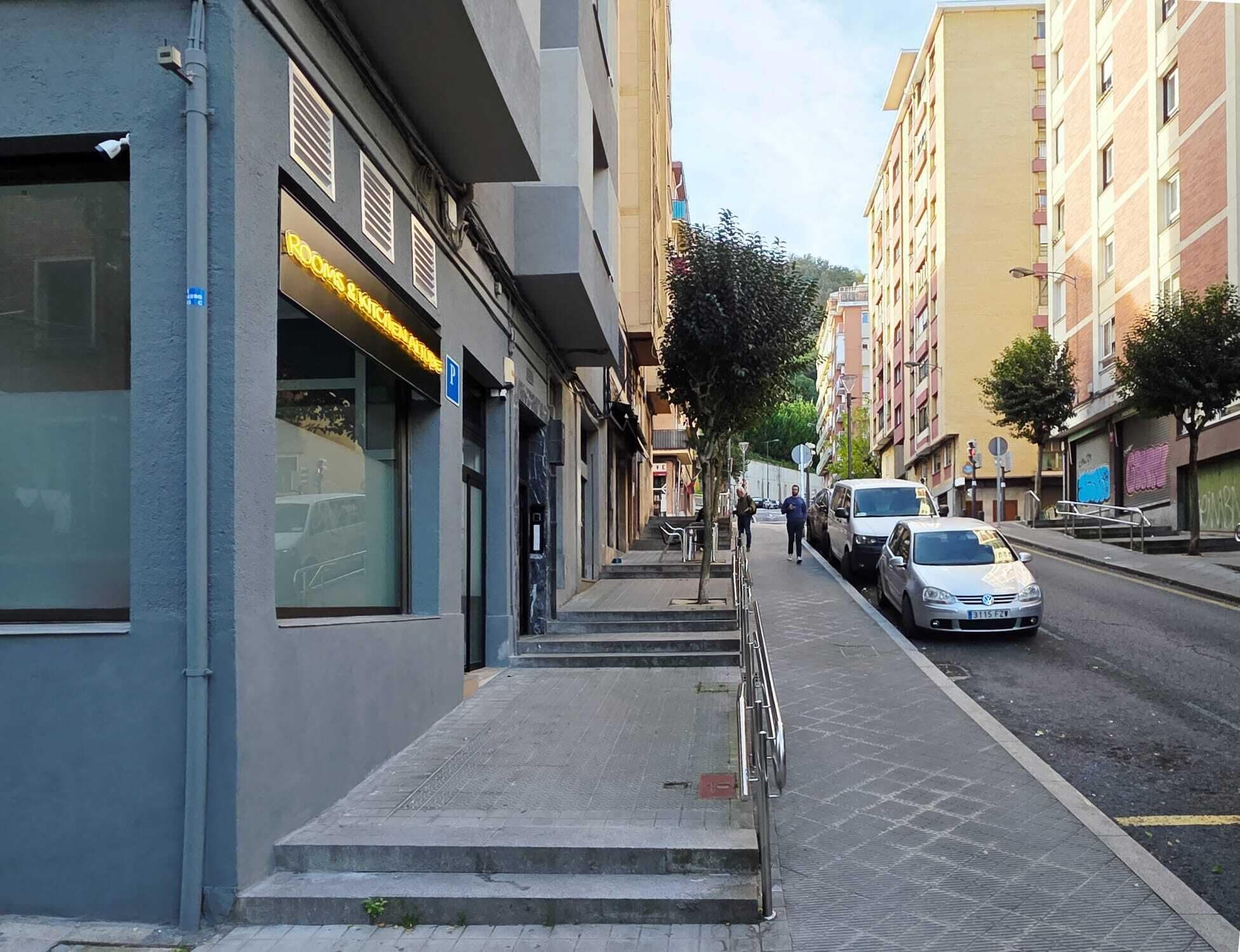 A street view with a building labeled 'ROCKS BIKES' on the left, a sidewalk, parked cars, and a pedestrian walking.