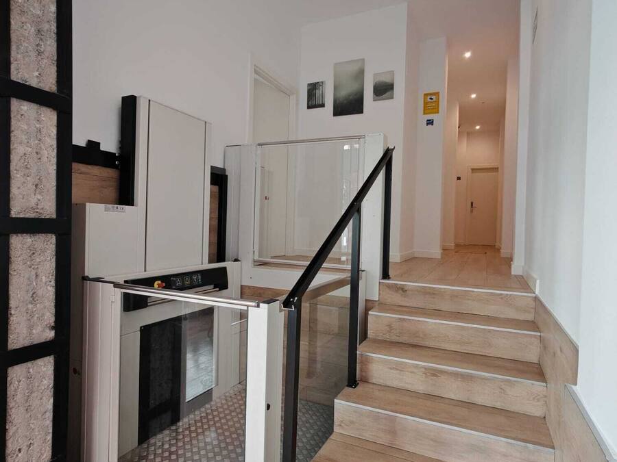 Una escalera conduce a una plataforma con barandilla de vidrio, una puerta blanca y vista a un pasillo con una puerta al final.