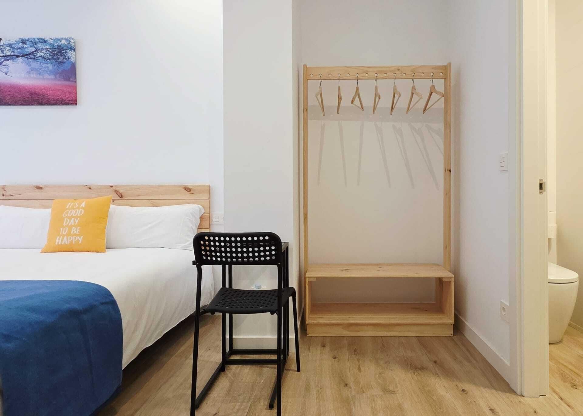 Un dormitorio con una cama, una silla, un perchero de madera y un pequeño banco.
