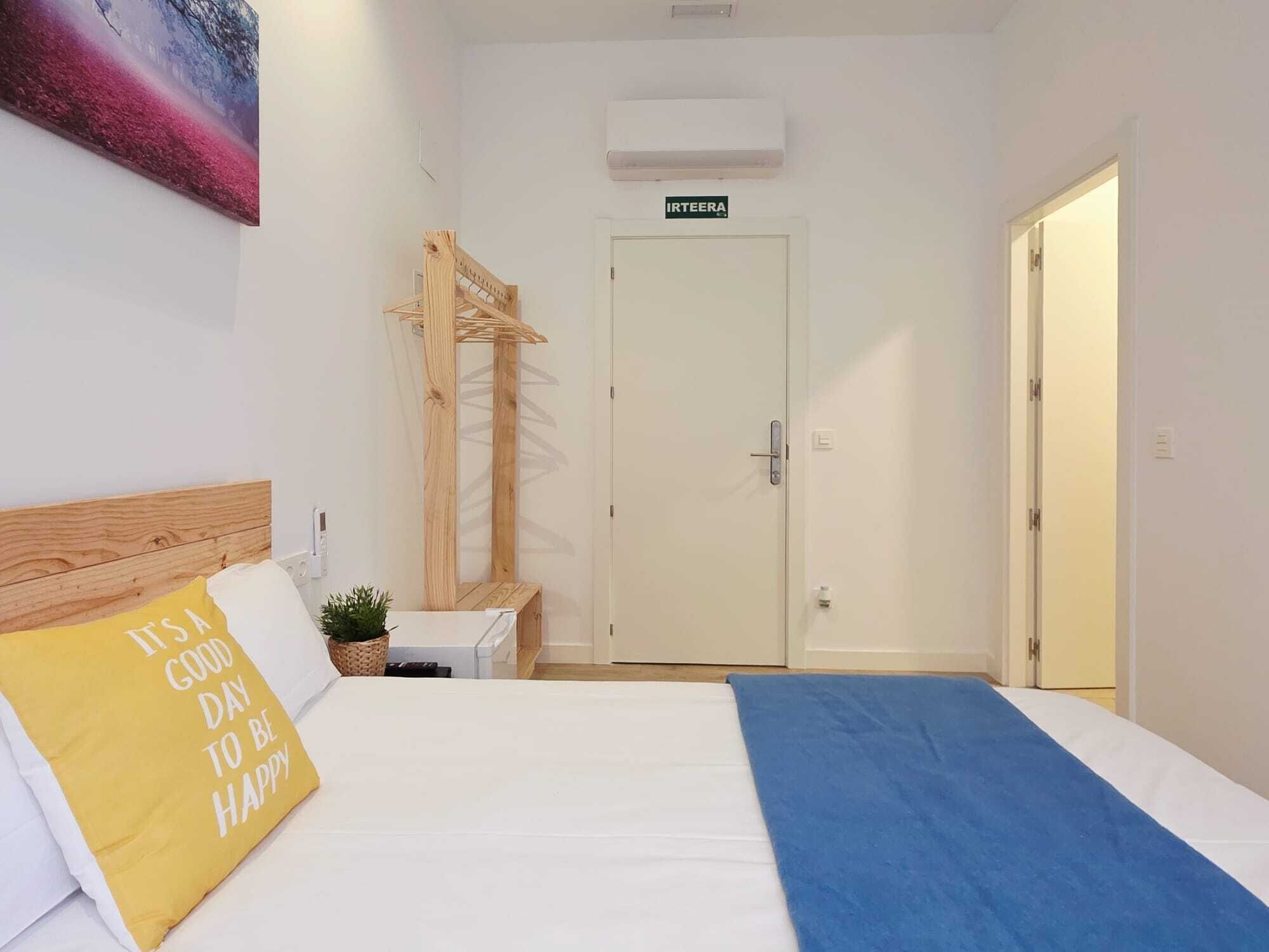 Habitación de hotel con cama, mesita de noche, aire acondicionado instalado en la pared, una puerta y un cartel sobre la puerta.